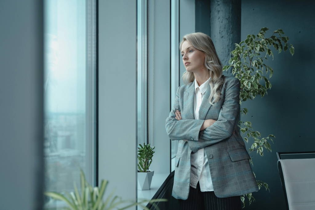 Professional woman engaging in self reflection while looking out office window, symbolizing leadership growth.