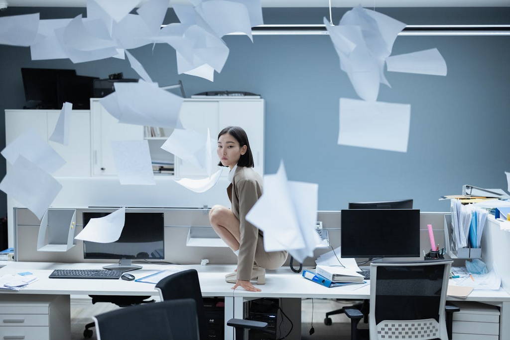 A woman crouched on an office desk with papers flying around, representing workplace chaos and the need for career change.